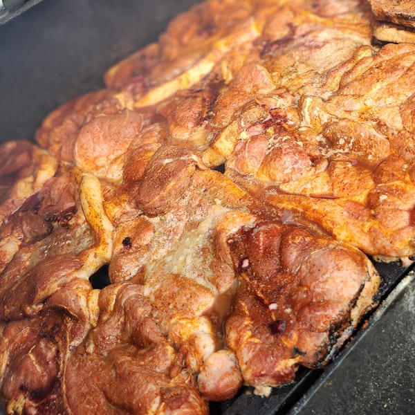 Close-up of seasoned pork shoulder steaks sizzling on a grill, with smoke rising.