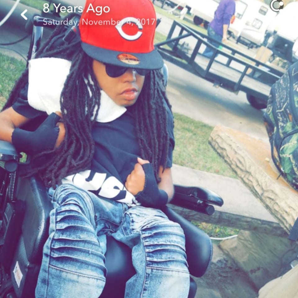 A person with dreadlocks, wearing a red baseball cap and sunglasses, sitting in a wheelchair outdoors.