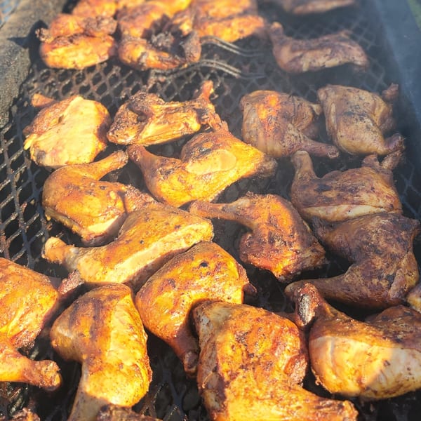 Close-up of numerous seasoned chicken pieces grilling on a dark metal grate, with some charring visible.